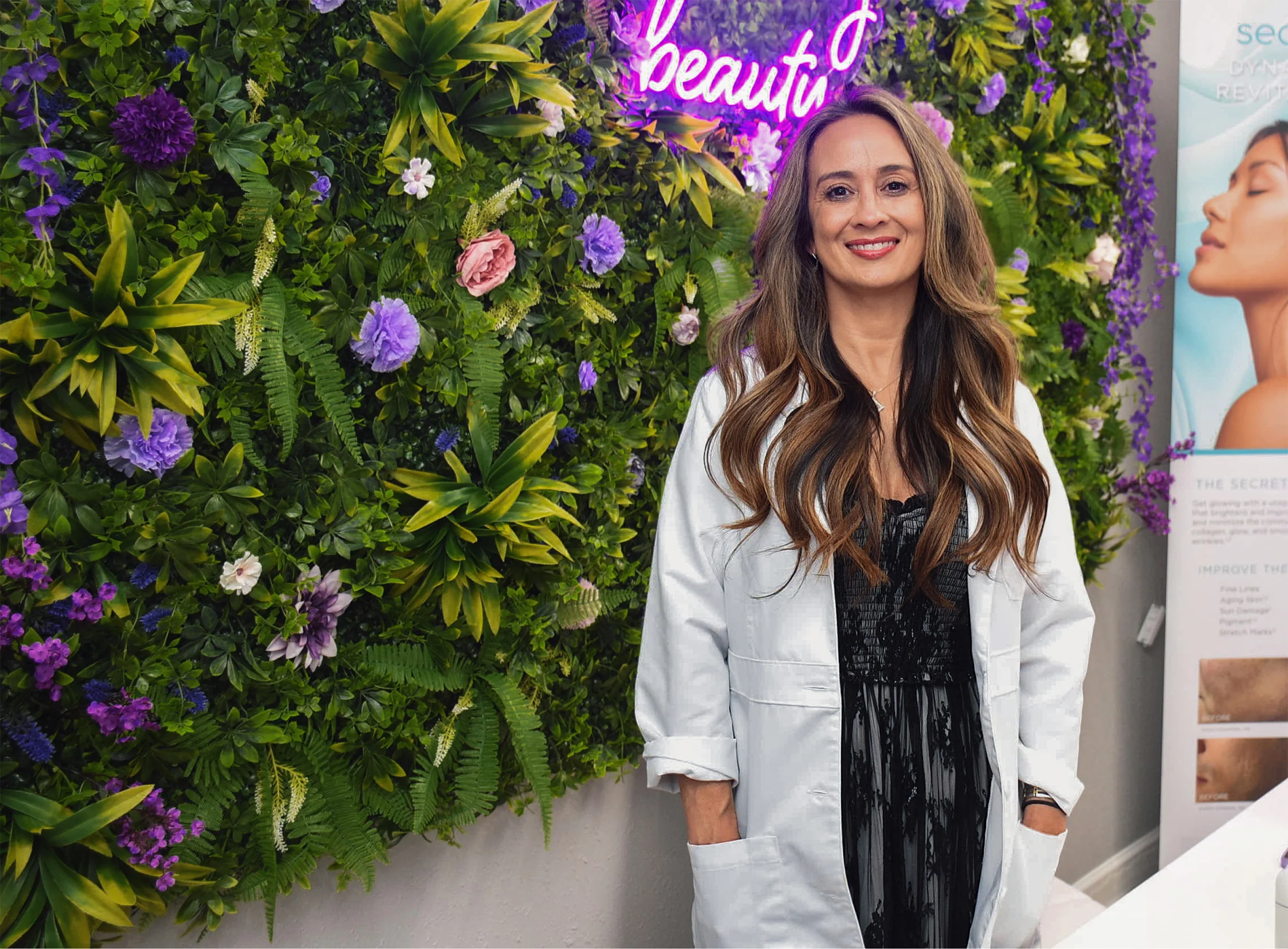 Woman in white lab coat smiling in front of floral wall with purple neon beauty sign - in Tampa, FL
