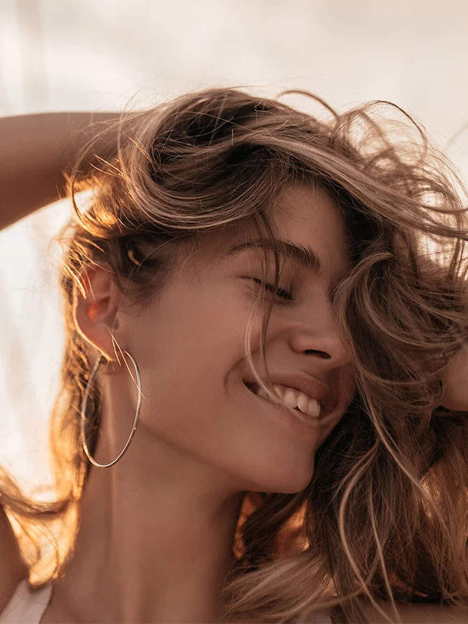 Woman with flowing brown hair smiling in golden sunlight wearing hoop earrings - IPL therapy in Tampa, FL