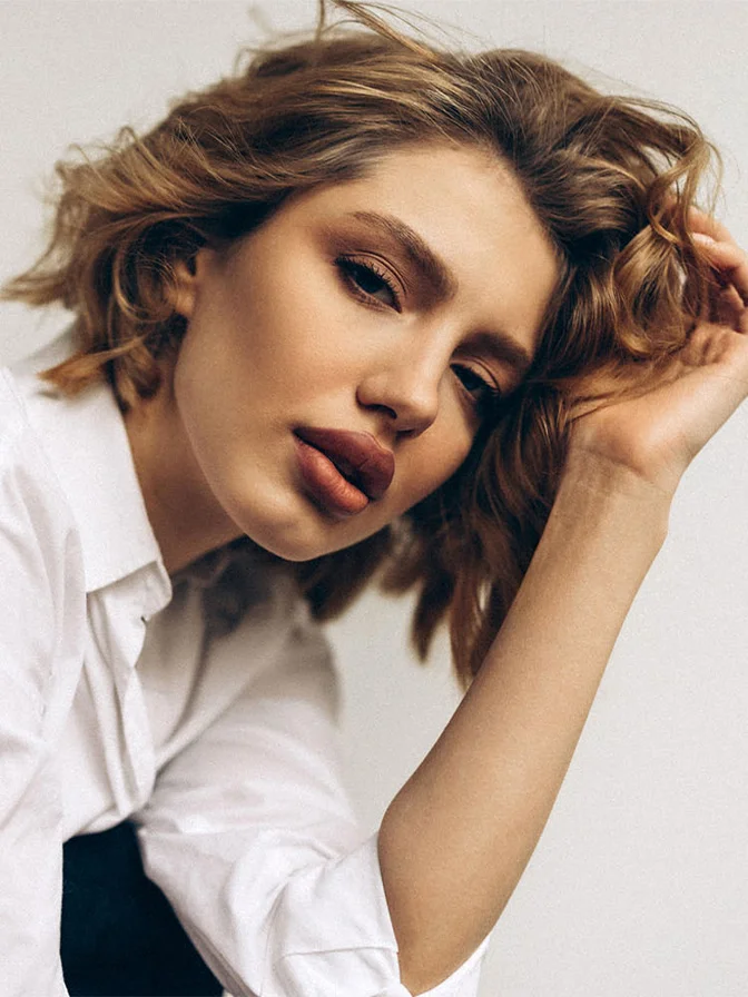 Woman with tousled wavy brown hair in white shirt posing with hand near face against neutral background - Liplase in Tampa, FL