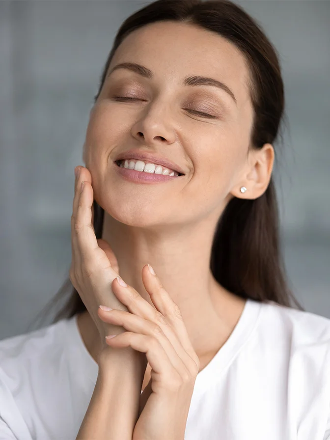 Woman with glowing skin touching face and smiling with eyes closed in white shirt against gray background - RF microneedling in Tampa, FL