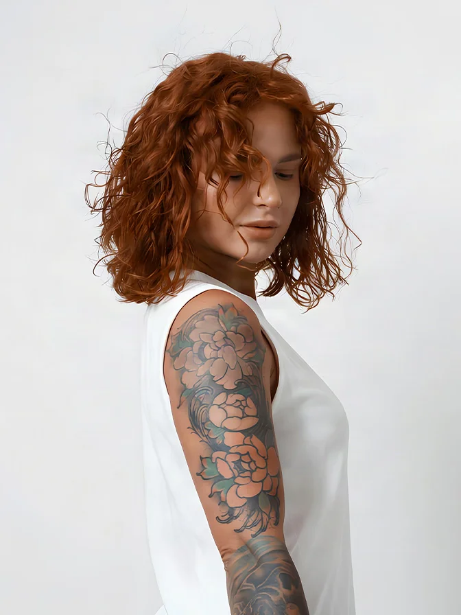 Person with curly red hair and floral sleeve tattoo wearing white tank top against gray background - Tattoo removal in Tampa, FL